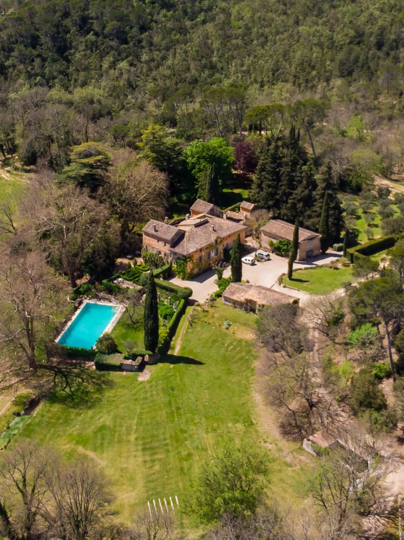 Aerial drone view of a large stone-built French country estate with terracotta rooftops, a rectangular turquoise swimming pool, manicured hedgerows, a gravel courtyard with parked vehicles, and surrounding woodland and green lawns.