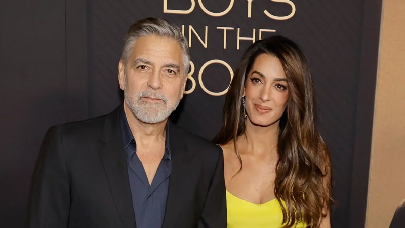 A man with gray hair and a short beard wearing a dark blazer over a navy shirt stands next to a woman with long brunette hair wearing a bright yellow dress, posing in front of a dark event backdrop.