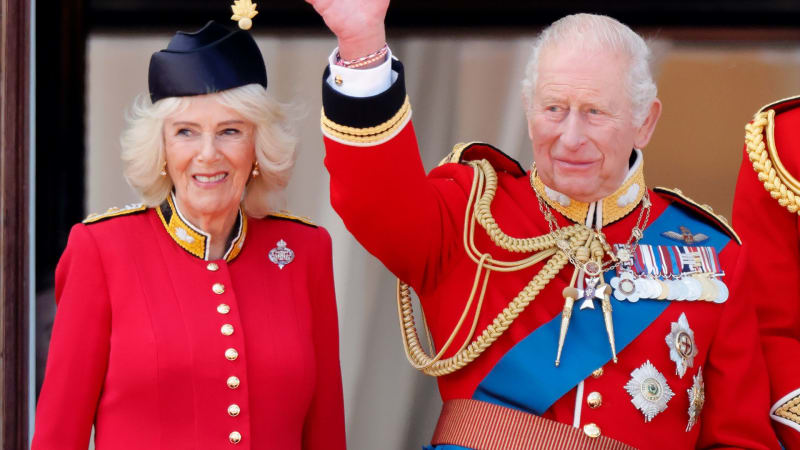 Two people in formal red ceremonial dress wave from a palace balcony during an official state occasion.