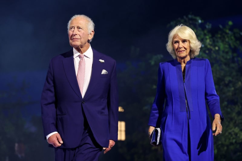Two people in formal blue attire walk together on a stage with a dark background and greenery visible behind them.