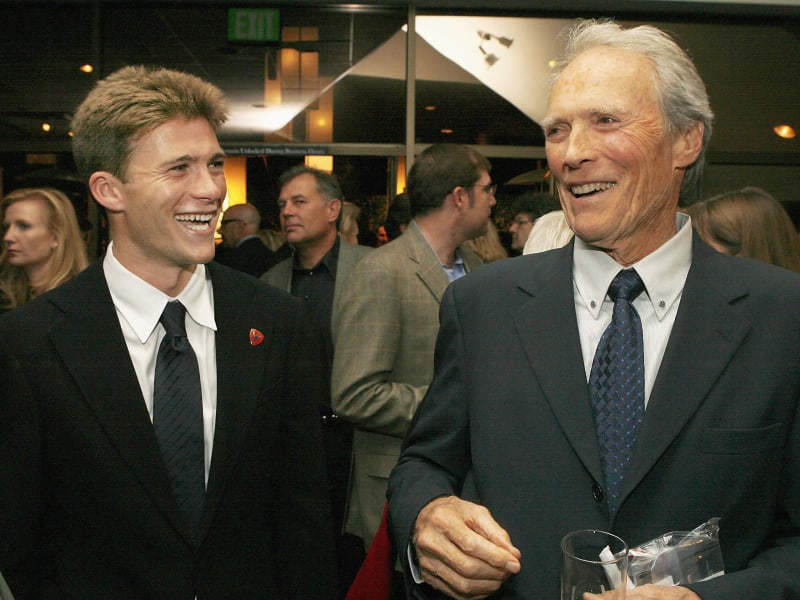 Two men in dark suits smile while conversing at a formal indoor event with other guests visible in the background.