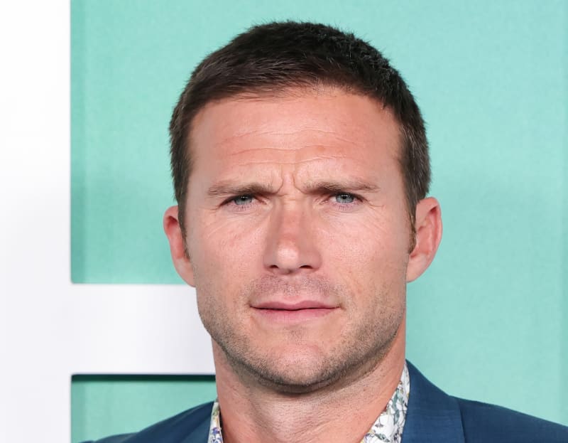 A man with short dark hair wearing a navy blue jacket stands against a turquoise and white geometric background.