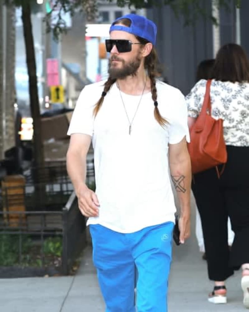 A man with long braided hair wears a blue cap, sunglasses, white t-shirt, and blue pants while walking on an urban street.