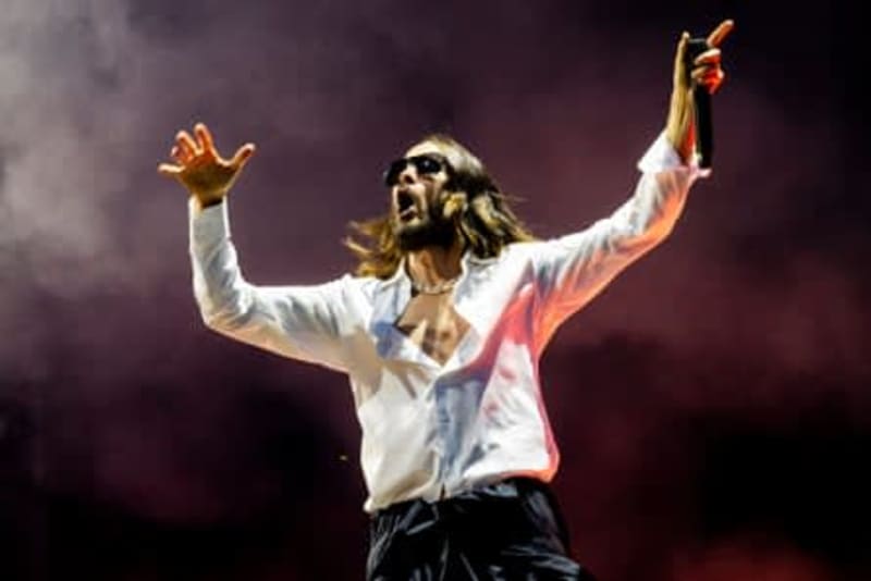 A performer with long hair wearing a white shirt and black pants stands on stage with arms raised in a victorious pose.