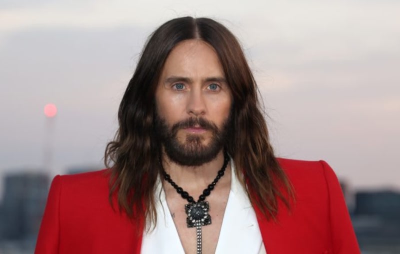 A man with long dark hair and a beard wearing a red blazer and white shirt with a pendant necklace stands outdoors at dusk.