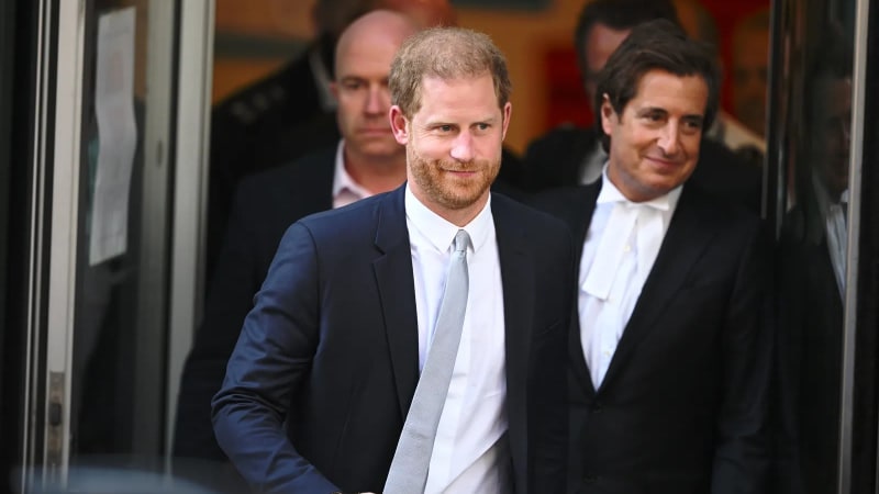 A red-bearded man in a navy suit and grey tie walks through glass doors, with a man in legal barrister's robes and bands visible to his right, and a third man in the background.