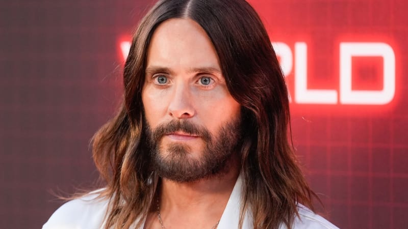 A man with long dark brown wavy hair, blue eyes, and a full beard, wearing a white open-collar shirt, posed in front of a red illuminated sign backdrop.