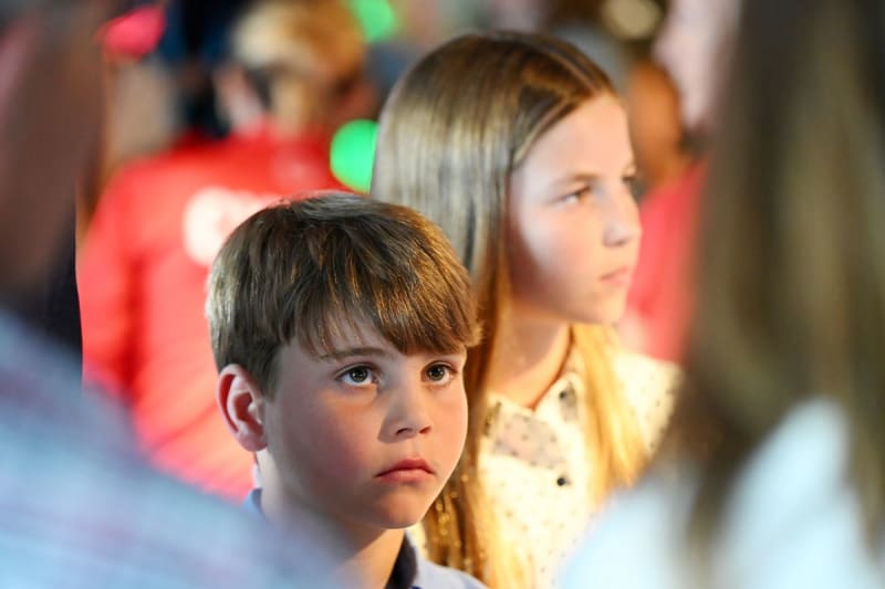 Two children standing attentively in a crowded indoor venue with festive red and green lights in the background.