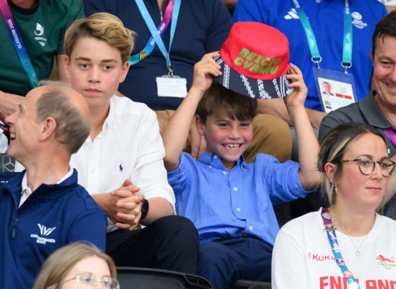 A young boy in a blue shirt holds up a red cap while sitting among spectators wearing Olympic lanyards at a sporting event.