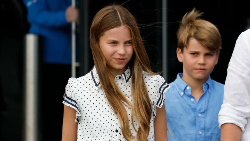 A girl with long brown hair wearing a white polka dot dress with navy trim stands in front of a boy with short light brown hair wearing a light blue button-up shirt, both standing outdoors with a dark blurred background.