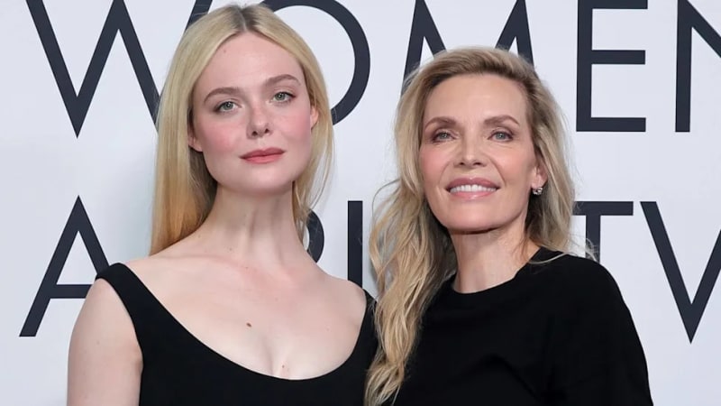 Two women in black attire pose together at a formal event against a white backdrop with partial text visible.