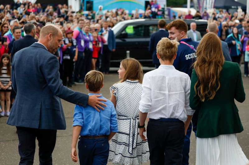 A man in a blue suit greets children at a public event with large crowds gathered in the background.