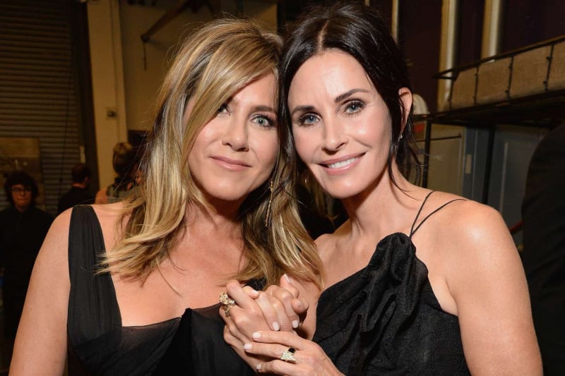 Two women in black dresses standing close together holding hands, posing for a photo indoors at what appears to be an event venue.