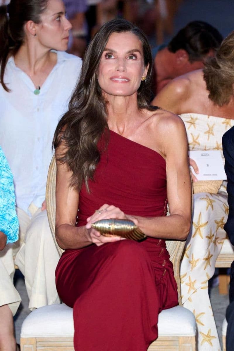 Woman in burgundy one-shoulder dress holding gold clutch seated at formal event among other guests.