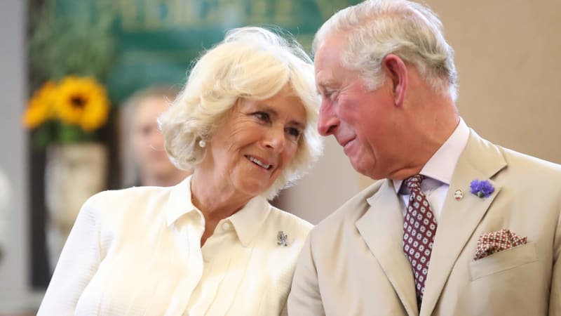 Two elegantly dressed older adults in cream-colored formal attire smile at each other at an indoor event with flowers in the background.