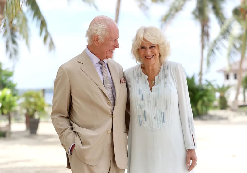 An older man in a tan suit and patterned tie stands beside an older woman in a light blue embroidered dress, both facing each other and smiling, with palm trees and a sandy beach setting in the background.