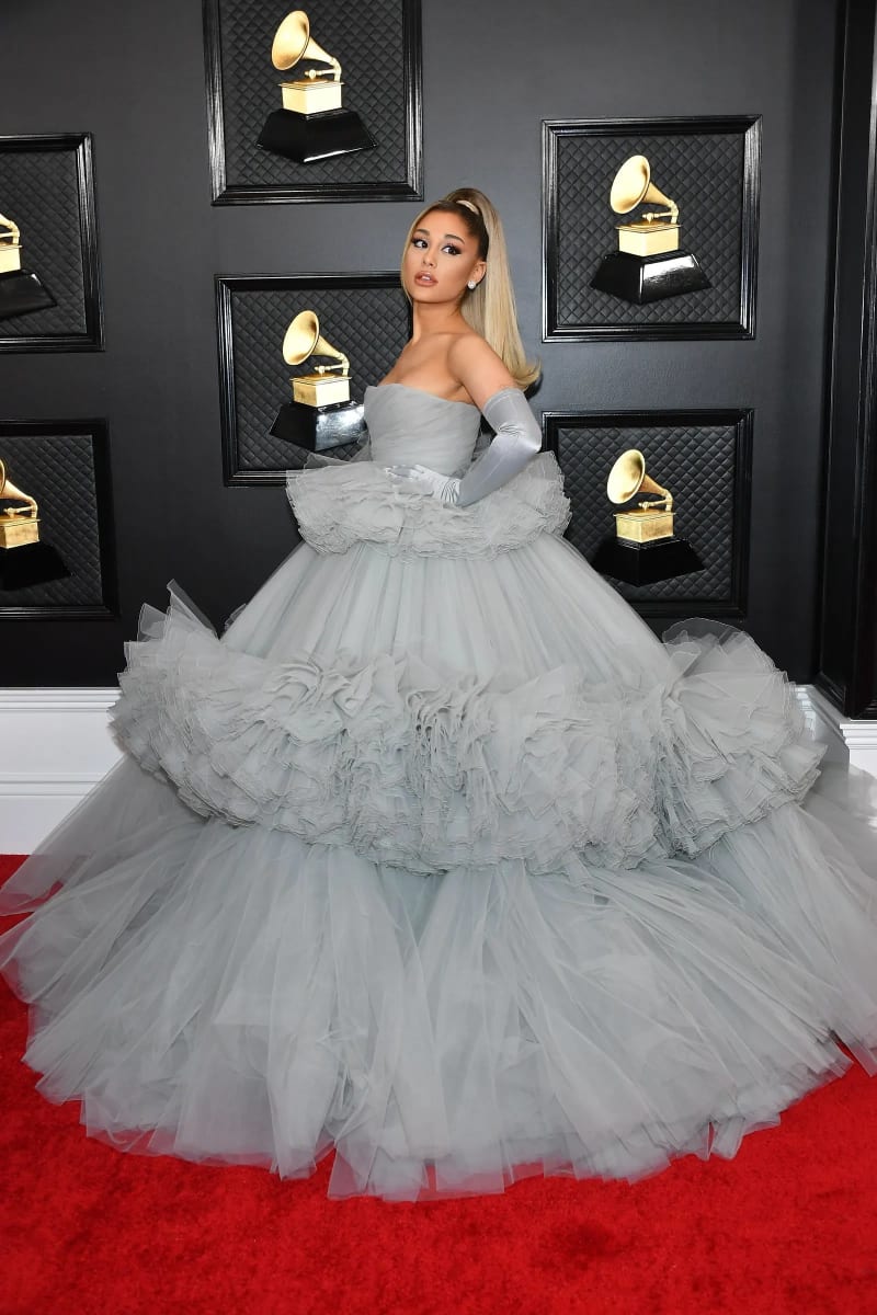 A woman with a long high ponytail wearing a strapless pale blue-grey tiered ruffle ballgown and matching elbow-length satin gloves, standing on a red carpet in front of a dark quilted backdrop displaying multiple Grammy Award trophies.