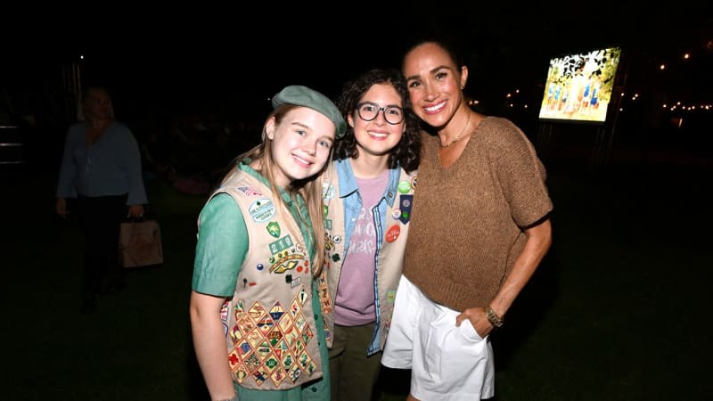 Three smiling people pose together at a nighttime outdoor event, with two wearing Girl Scout uniforms decorated with badges.