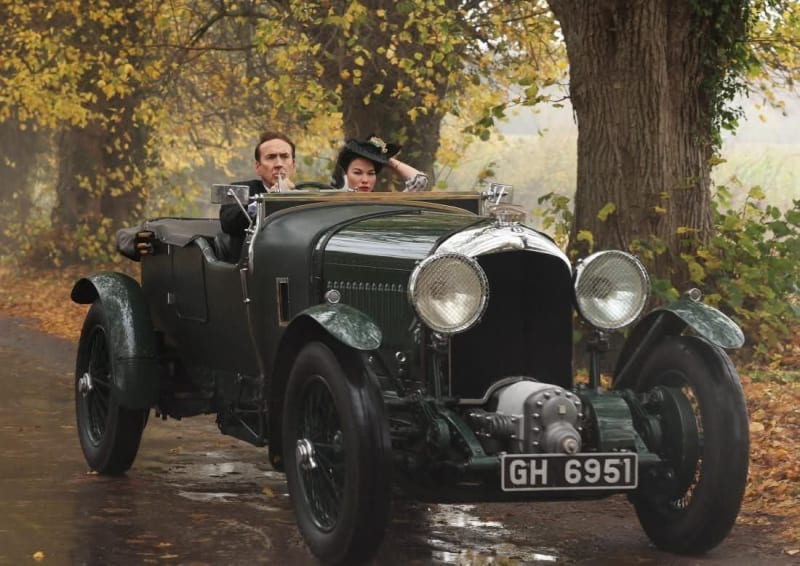 A man in a dark suit driving a vintage dark green open-top car with a woman in period costume and a decorated hat seated beside him, on a wet rural road surrounded by autumn trees.