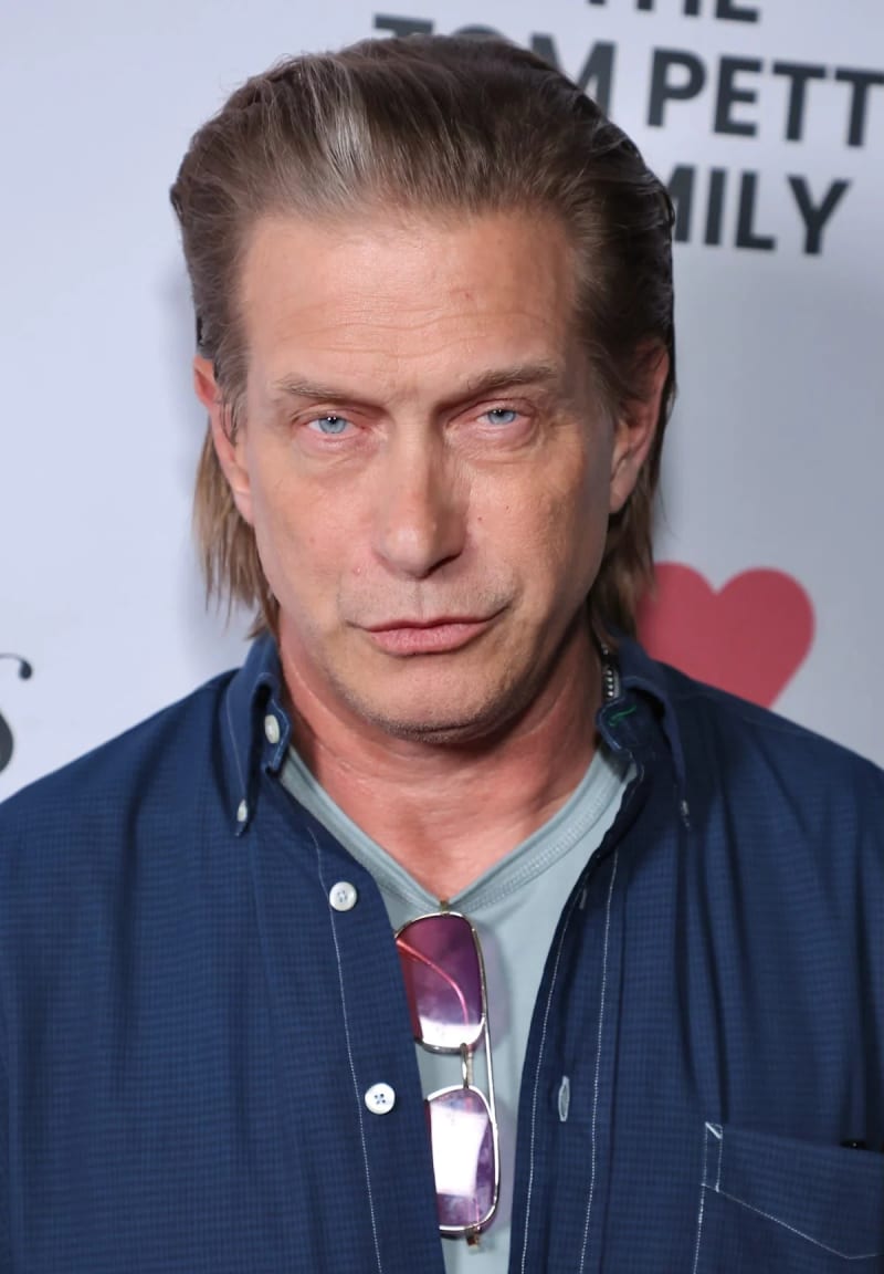 A middle-aged man with shoulder-length dirty blonde hair, blue eyes, wearing a blue button-down shirt over a grey t-shirt with sunglasses tucked into the collar, standing in front of a Tom Petty Family branded backdrop.