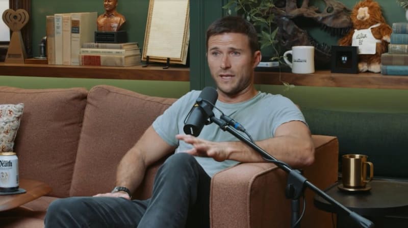 A man in a light blue t-shirt sits on a brown couch speaking into a microphone during what appears to be a podcast or interview.