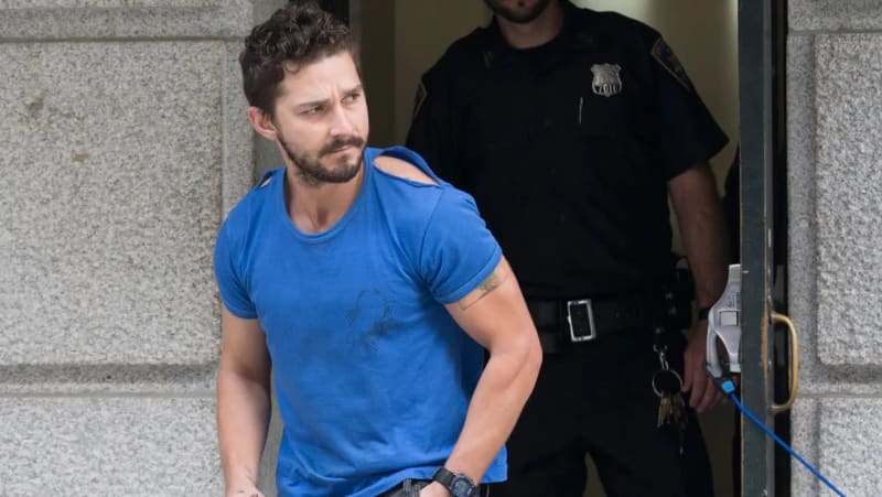 A bearded man with curly dark hair wearing a torn blue t-shirt walks out of a stone building doorway, with a uniformed police officer standing behind him.