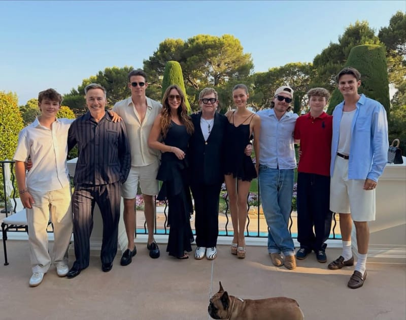 A group of ten people posing together on a sunny terrace with Mediterranean landscaping and a small dog in the foreground.