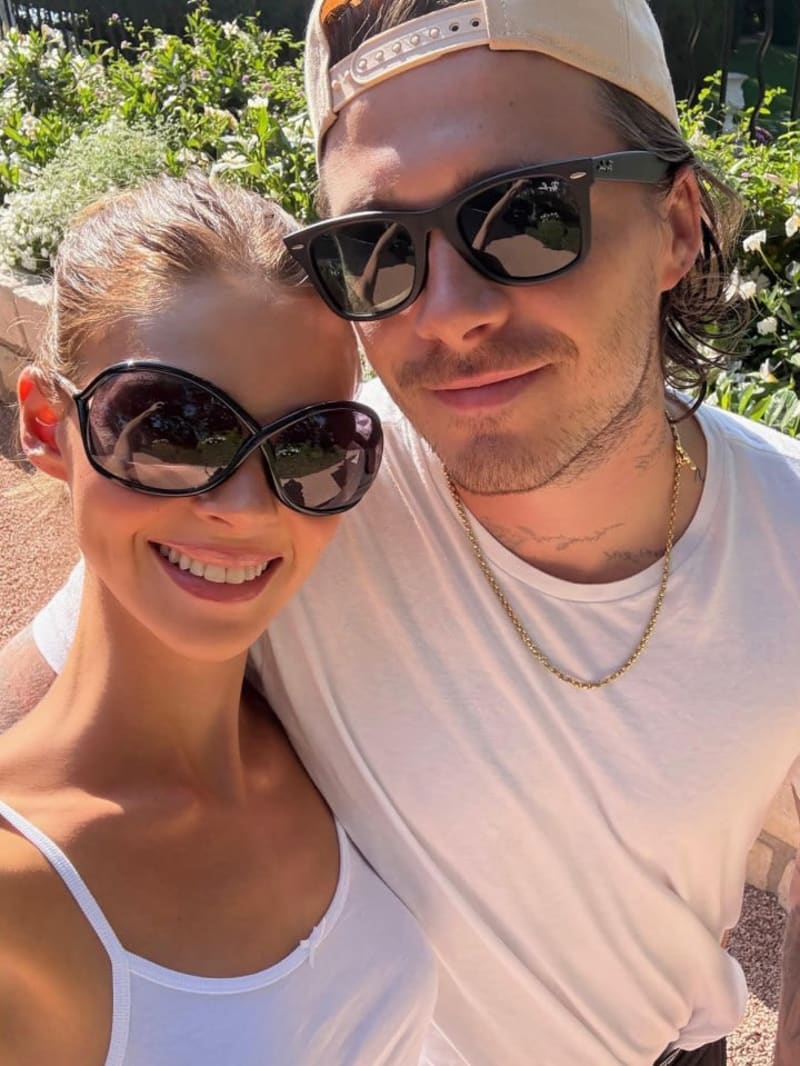 Two people wearing sunglasses and white shirts pose together outdoors on a sunny day with green foliage in the background.