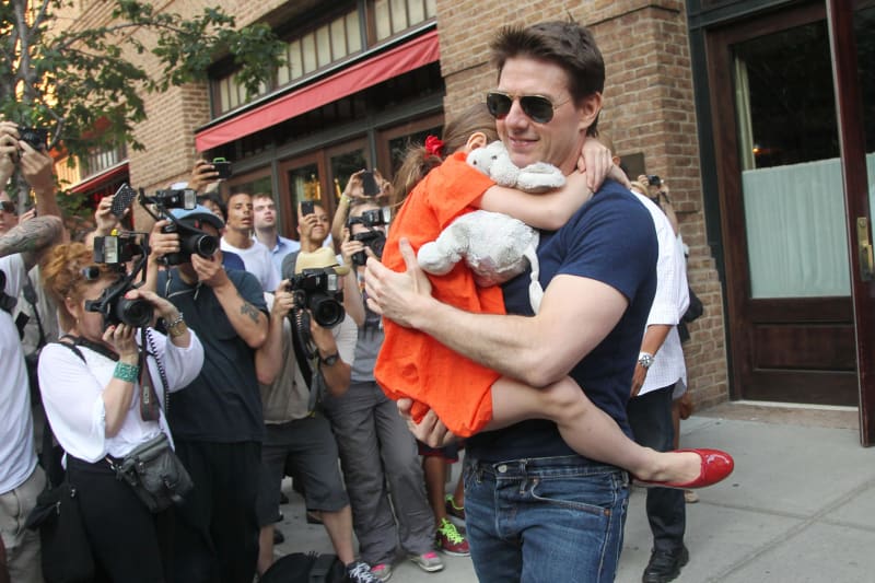 A man wearing aviator sunglasses, a navy t-shirt, and jeans carries a young girl dressed in an orange outfit and red shoes, holding a stuffed animal, on a city sidewalk surrounded by a crowd of photographers.