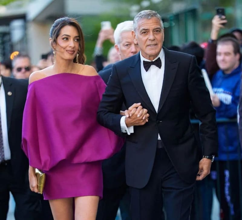 A couple in formal attire walks together at an elegant event, with the woman wearing a magenta off-shoulder dress and the man in a black tuxedo.