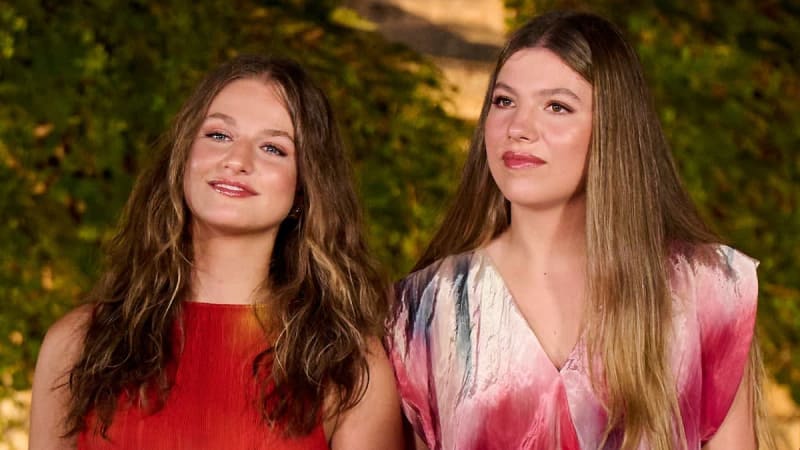 Two young women with long brown hair stand together against a green foliage background, wearing red and pink clothing.