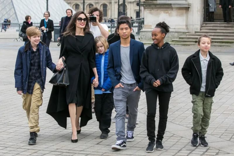 A woman in black sunglasses and coat walks with six children of various ages across a public plaza.