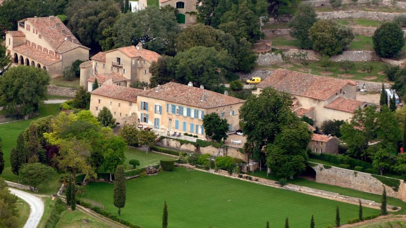 Aerial view of a large French stone château with terracotta roof tiles, blue shuttered windows, manicured green lawns, cypress trees, and surrounding countryside buildings.