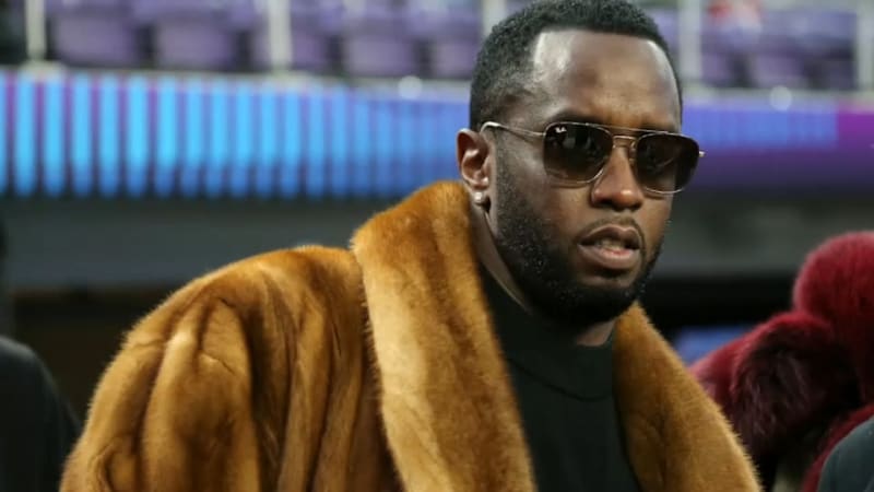 A man wearing aviator sunglasses, a black turtleneck, and a large tan fur coat stands in what appears to be a stadium with purple seats visible in the background.