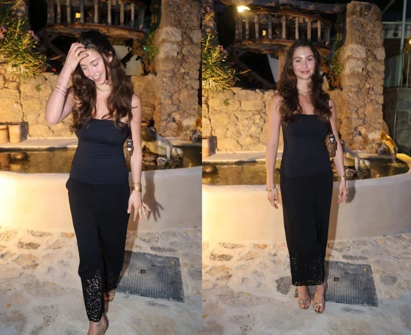 Two side-by-side photos of the same young woman with long wavy brown hair wearing a black strapless top, black laser-cut wide-leg trousers, gold strappy heels, and gold jewelry, standing outdoors at night in front of a stone fountain and rustic wooden structure, laughing with her hand in her hair in the left panel and posing directly for the camera in the right panel.