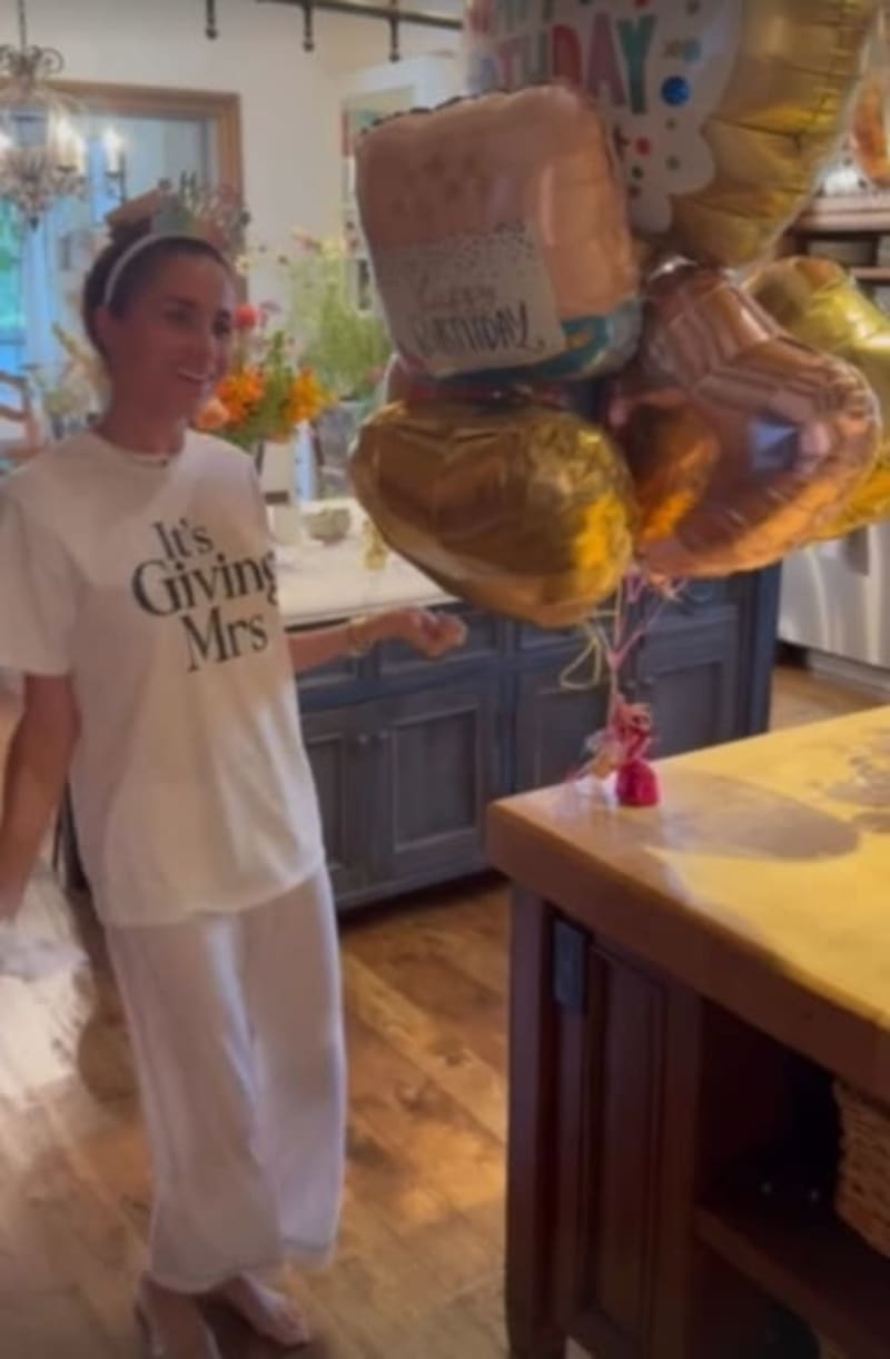 A woman wearing a "It's Giving Mrs" shirt stands in a kitchen beside gold and copper balloons.
