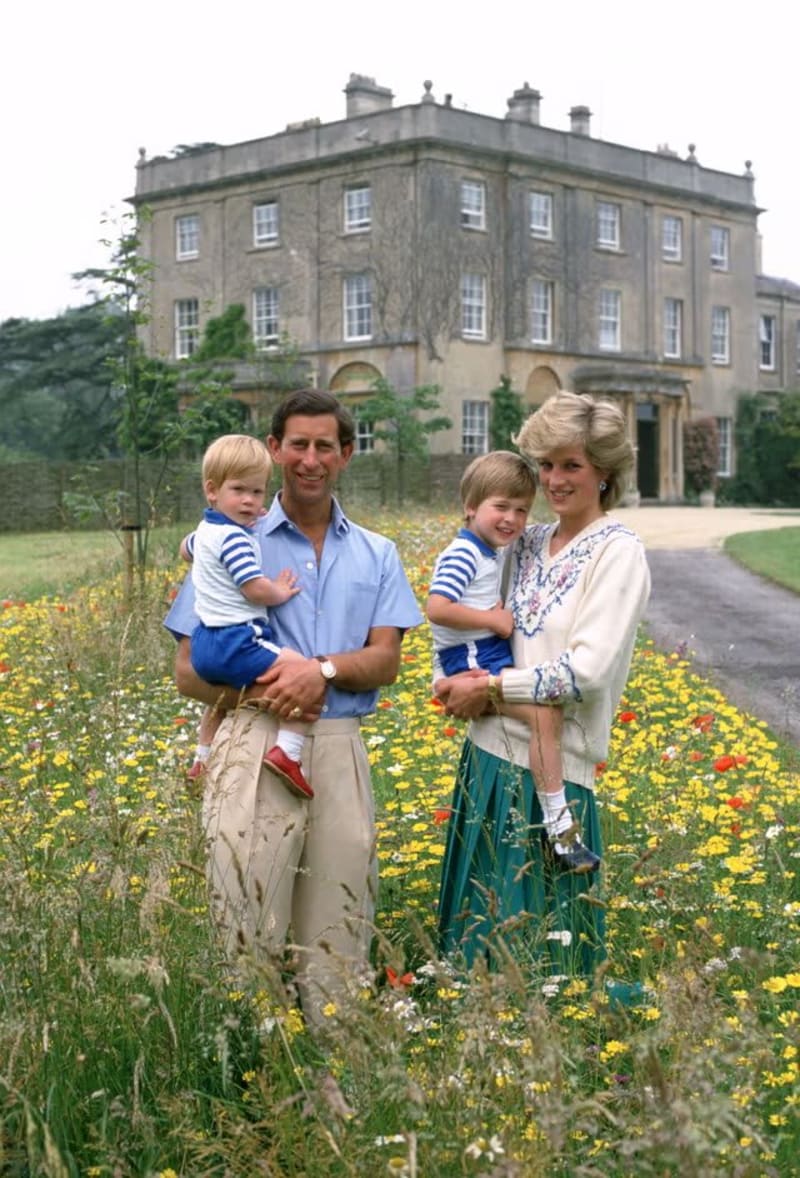 A man in a light blue short-sleeved shirt and khaki trousers holds a toddler in a striped top, standing beside a woman in a white floral blouse and teal skirt who holds a young boy in a striped shirt, all posing in a wildflower meadow in front of a large stone country house.
