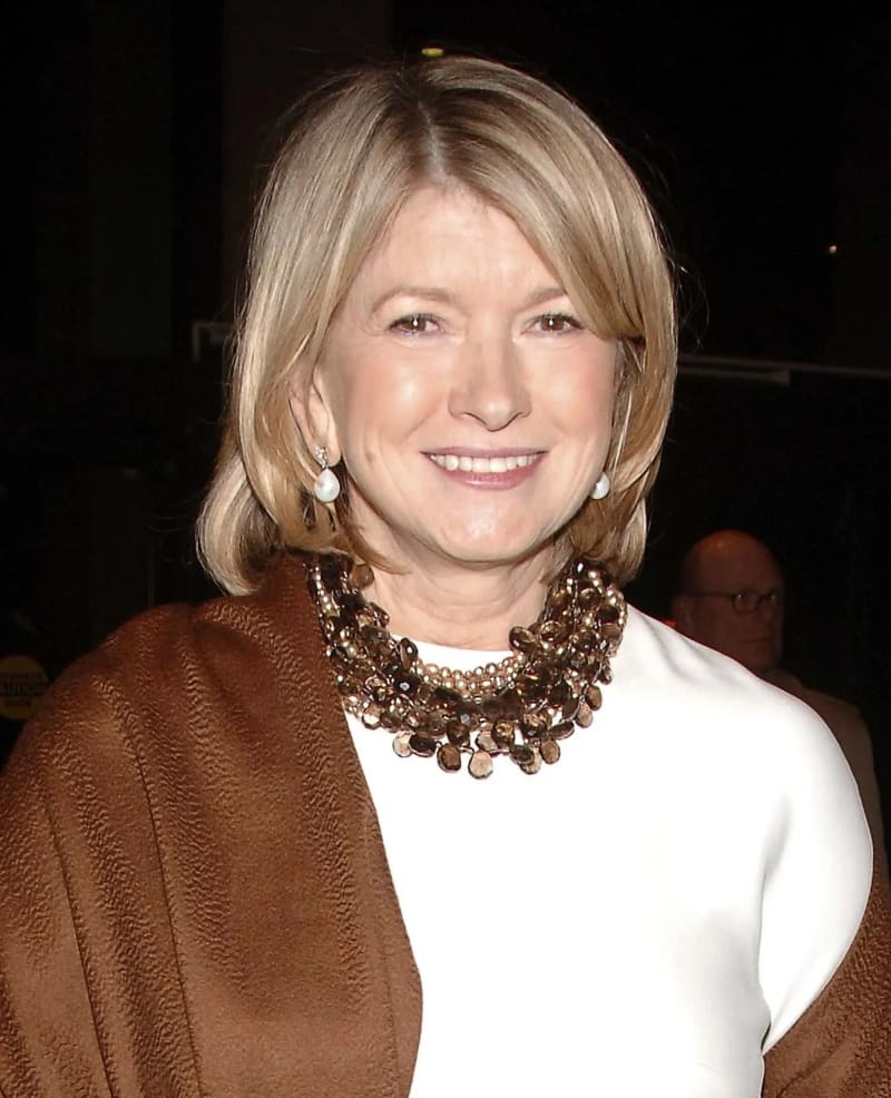 A woman with short blonde hair wearing a white top, a brown wrap, pearl drop earrings, and a layered bronze beaded necklace, smiling at an event against a dark background.