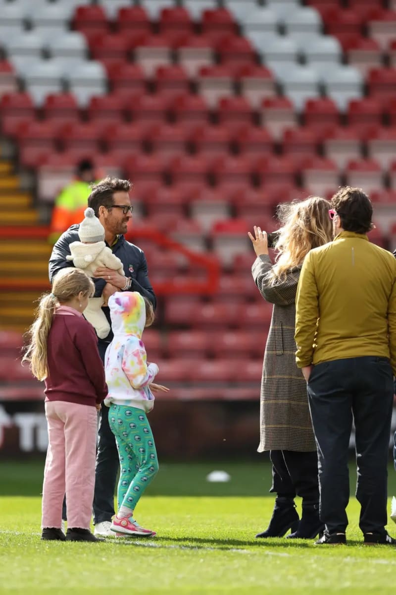 A man wearing glasses and a dark jacket holds a baby dressed in a white knit hat and cream outfit while standing on a grass football pitch with two young girls nearby; to his right, a woman in a plaid coat photographs the group alongside another person in a yellow jacket, with empty red stadium seats visible in the background.