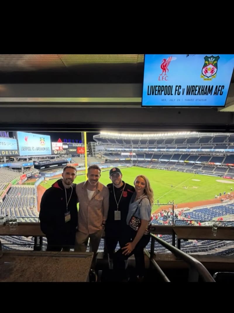 Four people — three men and a woman with long blonde hair — posing together in a stadium suite with a soccer pitch and a screen displaying 'Liverpool FC v Wrexham AFC' visible behind them.