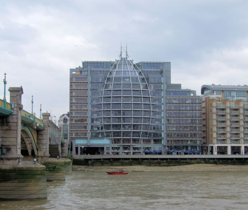 A large glass and steel office building with a distinctive oval-shaped lattice facade on the South Bank of the River Thames, with Southwark Bridge visible to the left and a small red boat on the river in the foreground, under an overcast sky.