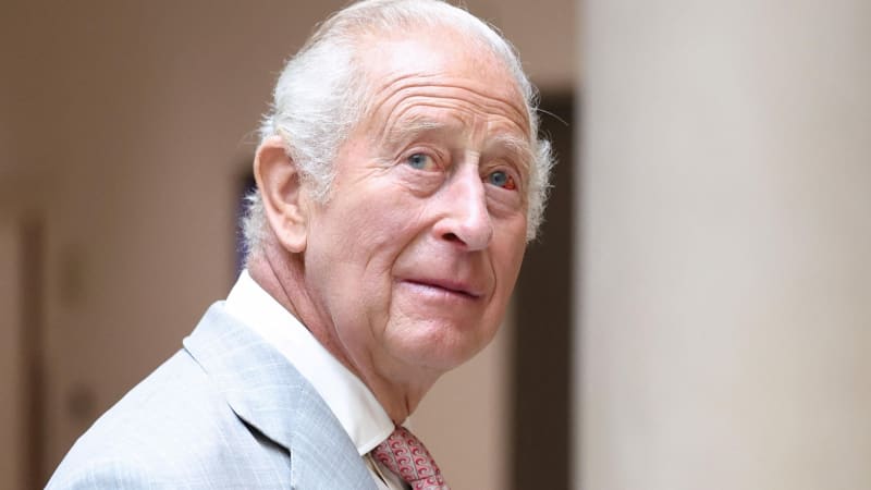 An elderly man with white hair wearing a light blue suit jacket, white dress shirt, and red patterned tie, photographed from the shoulders up against a blurred neutral indoor backdrop.
