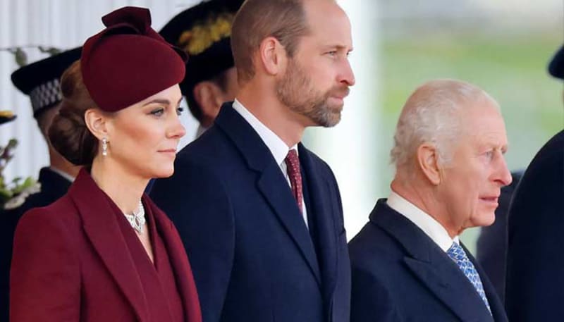 Three members of the British Royal Family stand together at an official outdoor event, dressed formally in dark suits and burgundy attire.