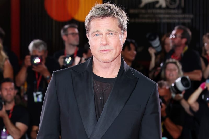 A man with short salt-and-pepper hair wearing a black blazer over a black t-shirt and a pendant necklace, standing in front of a crowd of photographers at a red carpet event.