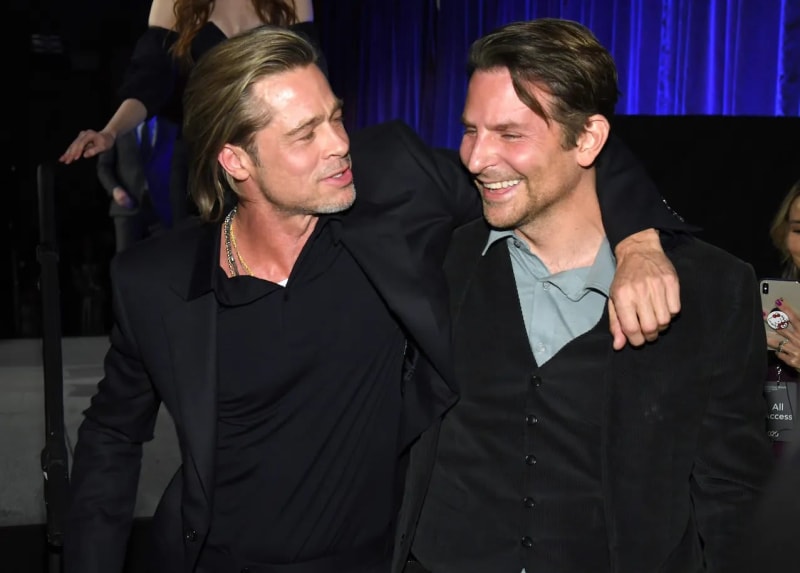 Two men in dark blazers stand together at an indoor event, with the man on the left draping his arm around the man on the right, who is laughing broadly, against a blue-lit backdrop.