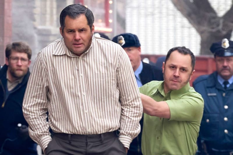 A man in a beige striped button-up shirt stands with hands behind his back while a man in a green polo shirt grips his arm from behind, with police officers and bystanders visible in the background on an urban street.