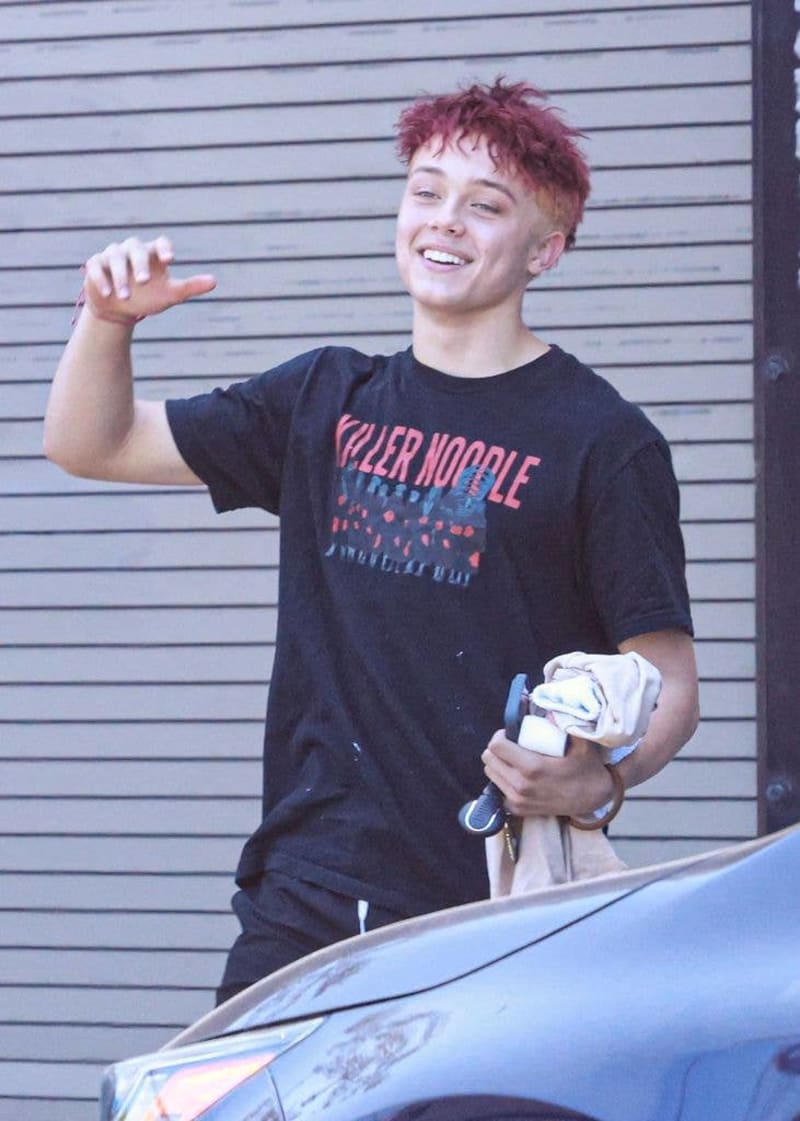 A smiling person with pink hair wearing a black "Roller Noodle" graphic t-shirt stands against a corrugated metal wall.