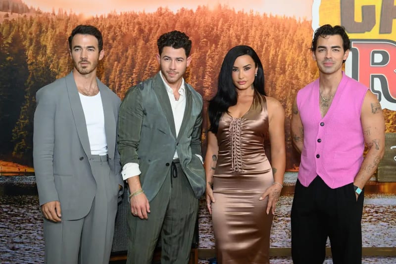 Four people standing together in front of a forest-themed backdrop, from left to right: a man in a grey suit with a white t-shirt, a man in a green velvet suit with an open white shirt, a woman with long black hair in a bronze satin halter dress, and a man in a pink sleeveless knit vest with black trousers.