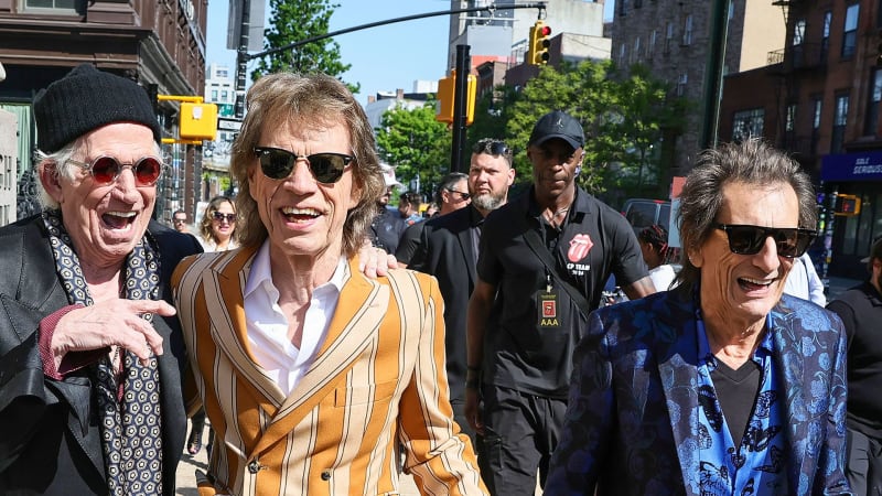 A group of elderly men in colorful suits and sunglasses smile while walking together on a city street lined with buildings.