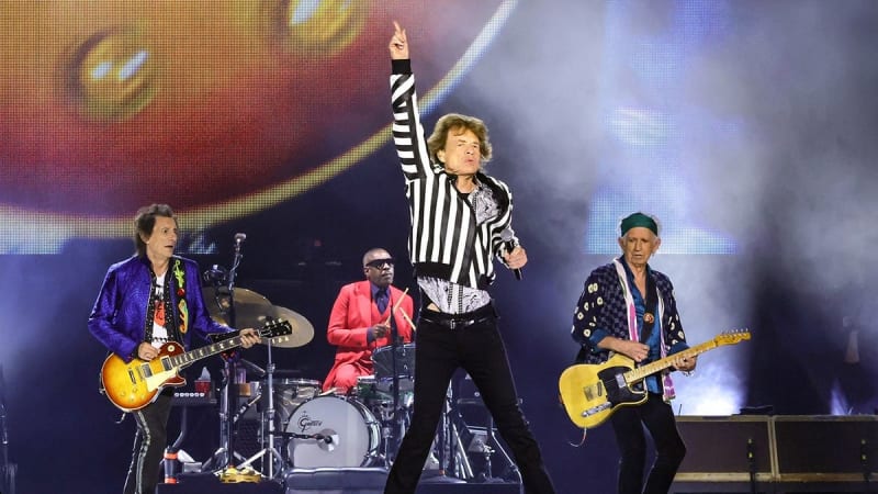 A rock band performs on stage with a lead singer in a striped jacket, guitarists, and drummer under colorful stage lighting.